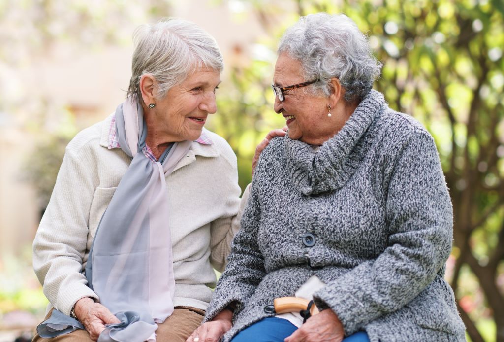 Two older adult woman sit together outside at an assisted living facility