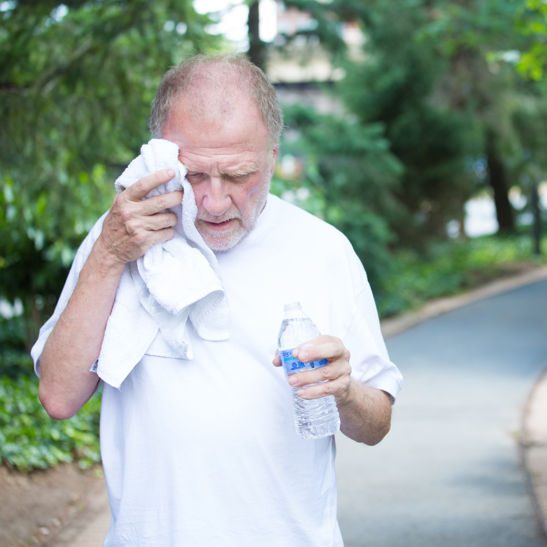 Elderly gentleman wearing a white teeshirt. He is on a walk on a hot day and has a towel that he's wiping his face with while holding a bottle of water.
