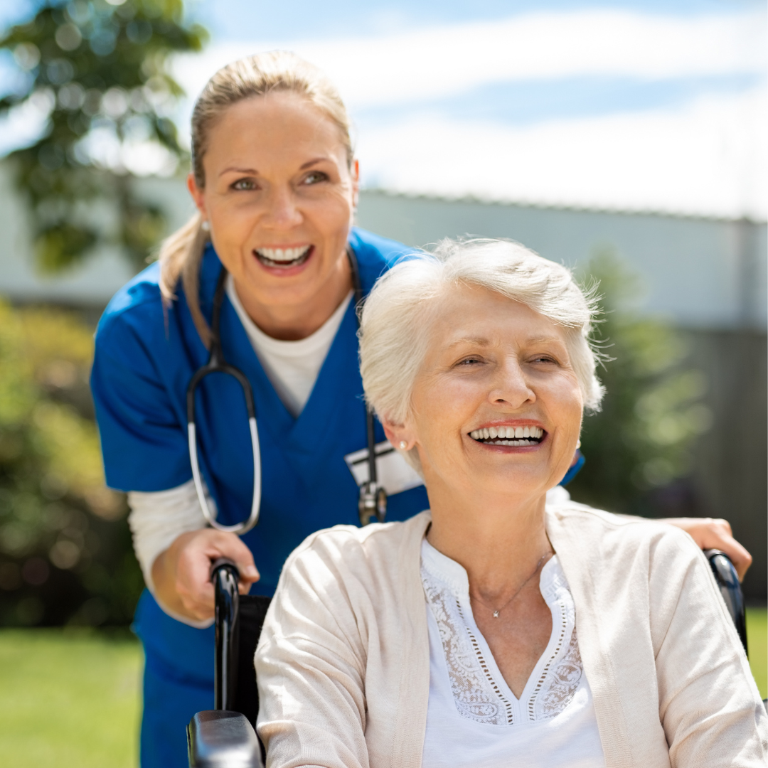 An older white haired woman is being pushed in a wheelchair by a woman in scrubs. They are both laughing and seem to be having a good time.