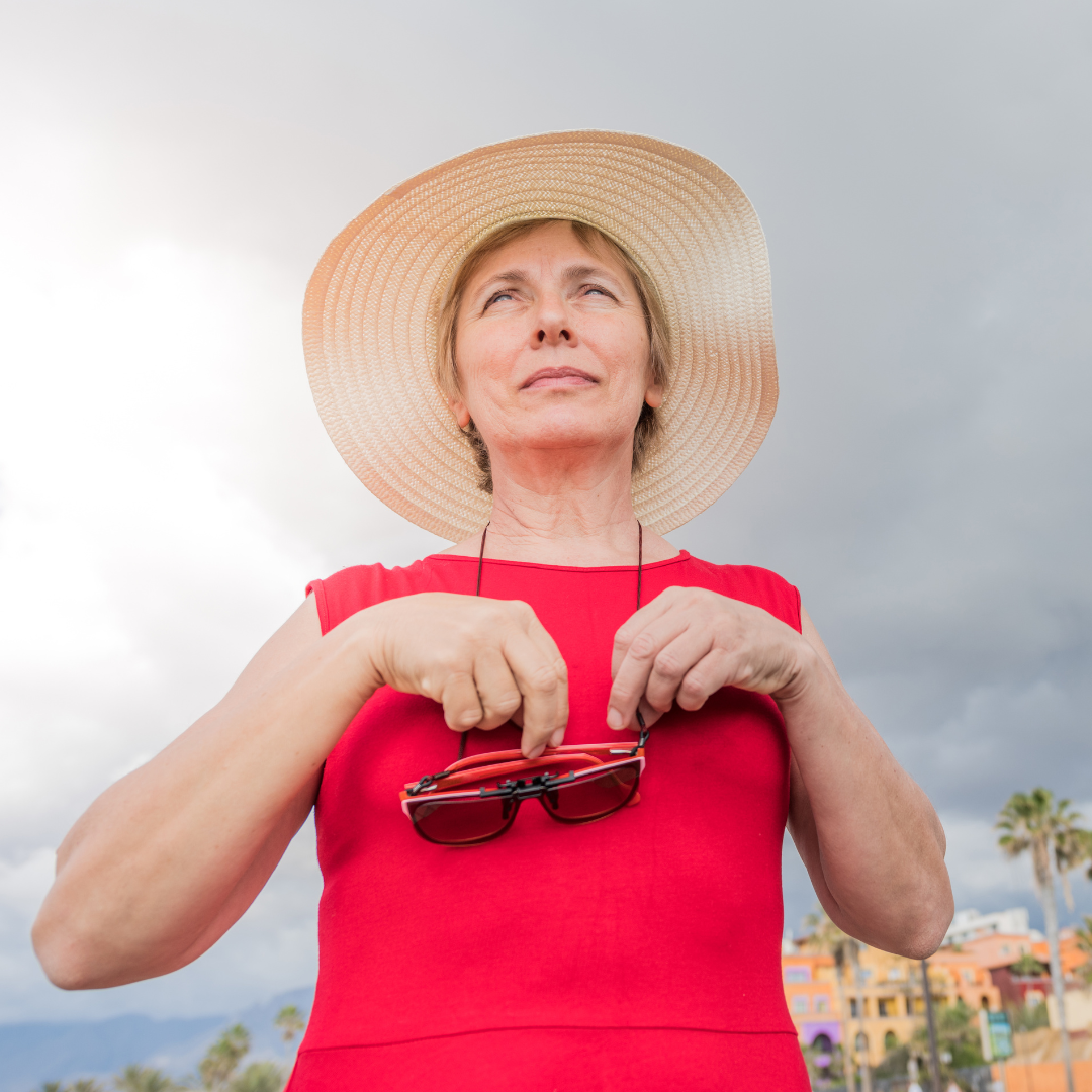 Photo of an older woman in a red dress wearing a large sun hat and holding a pair of sunglasses