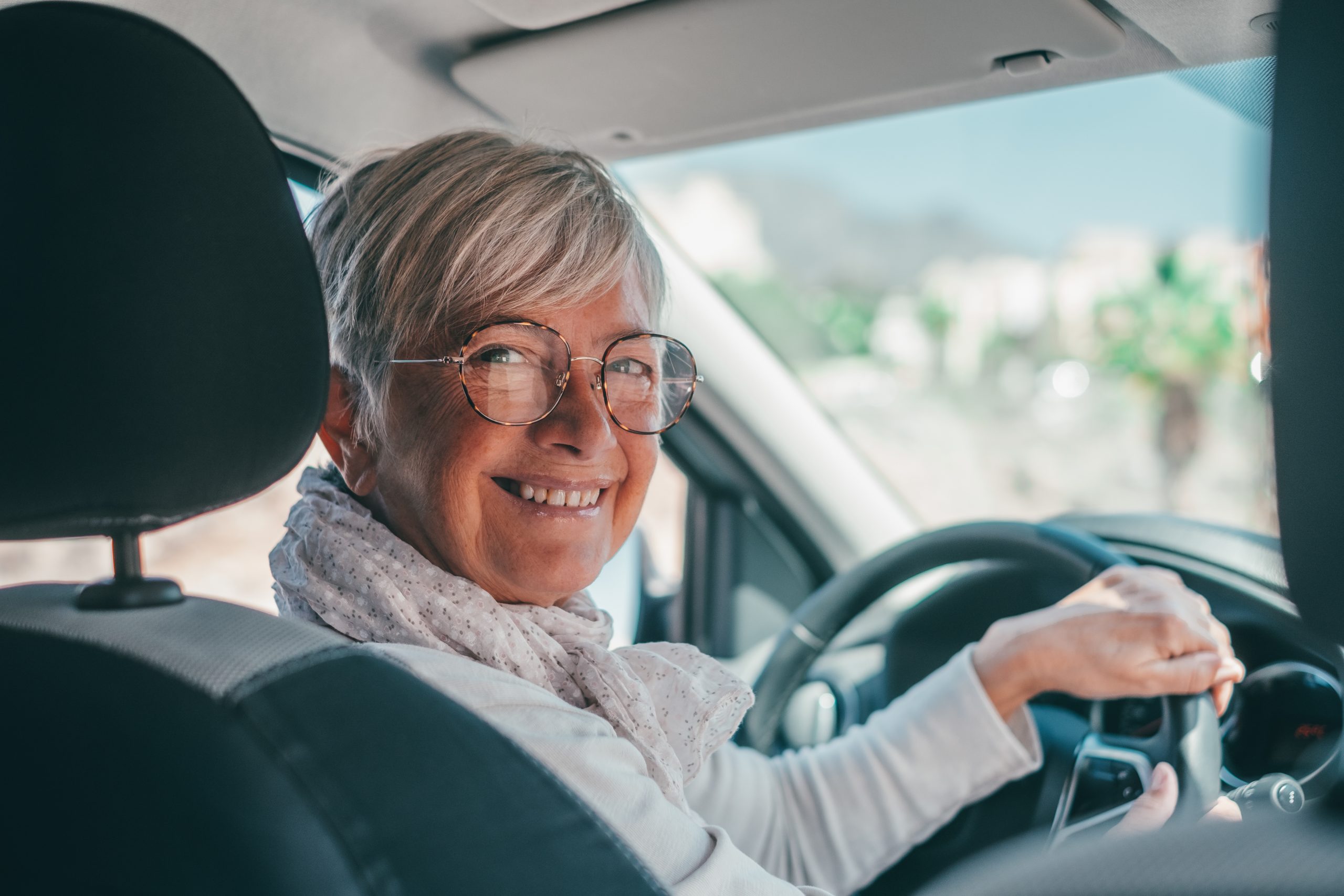 A gray-haired woman driving a car. She's staring back at the camera in the backseat smiling