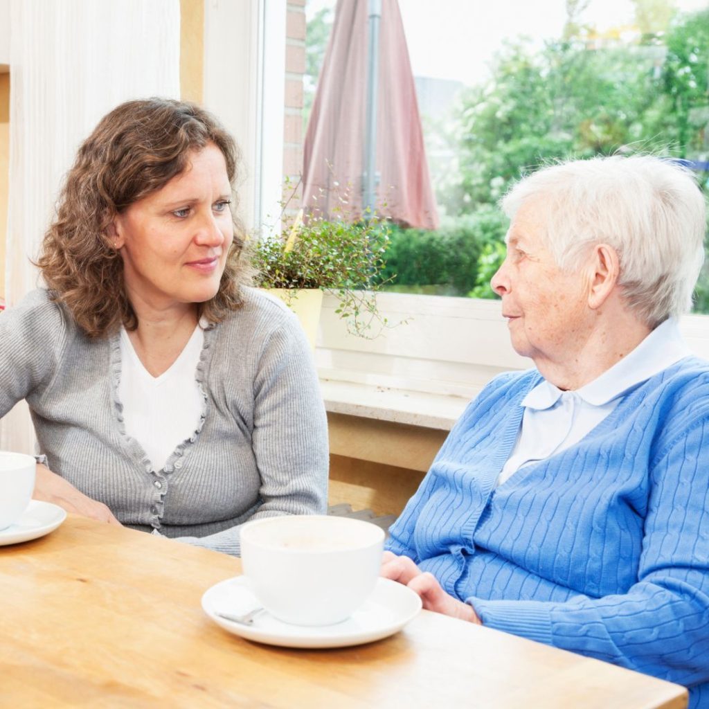 A woman in a gray cardigan listens to an elderly woman in a blue sweater over coffee.