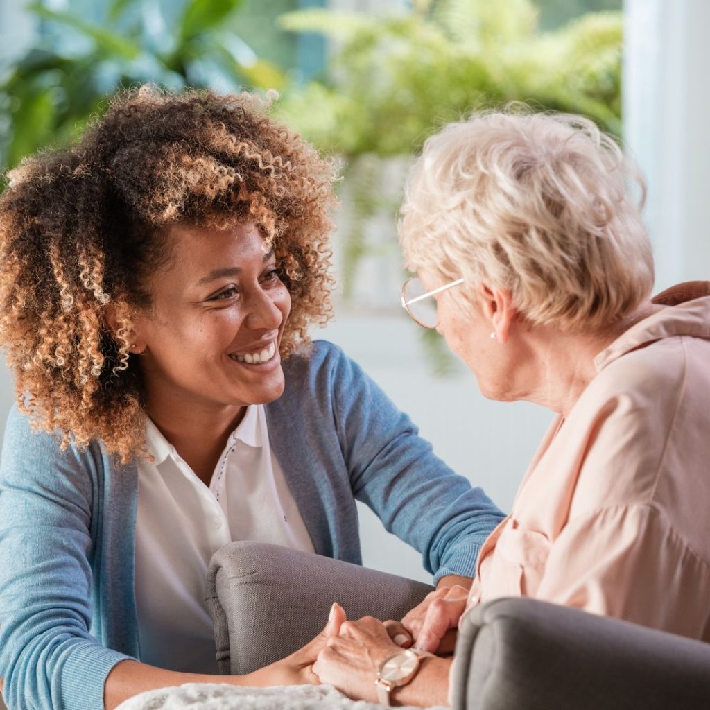 A woman in a blue cardigan is speaking to an older woman in a chair. The older woman's face is not visible. 