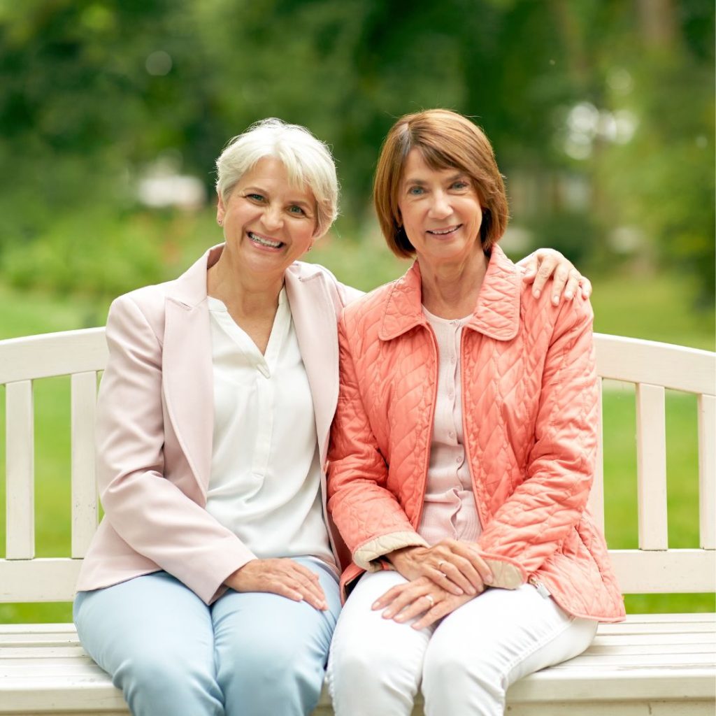 Two smiling older women sit closely on a white bench outdoors.