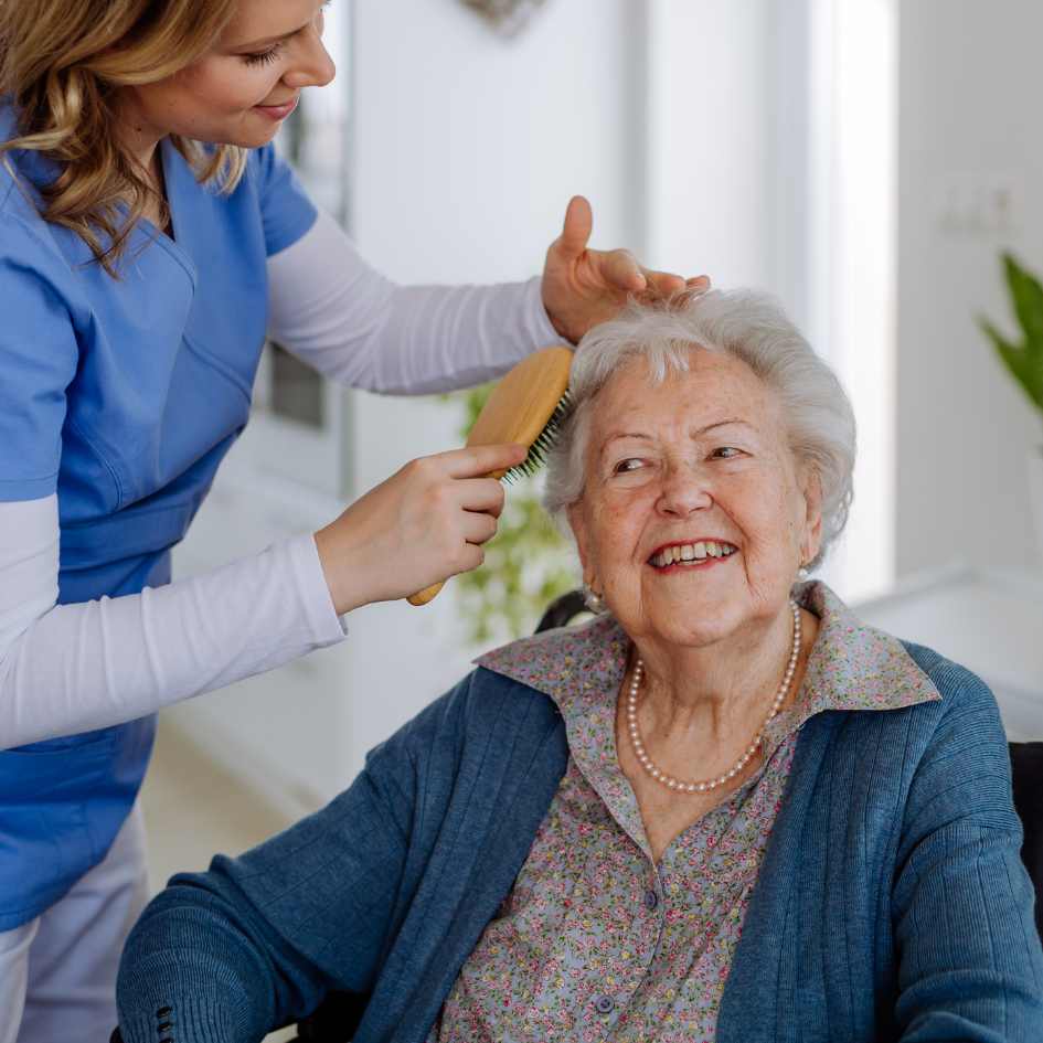 A woman helps an elderly woman who's sitting down brush her hair. 