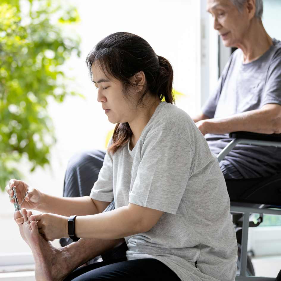 A young woman trims an elderly man's toenails as he sits in a wheelchair, with greenery in the background.