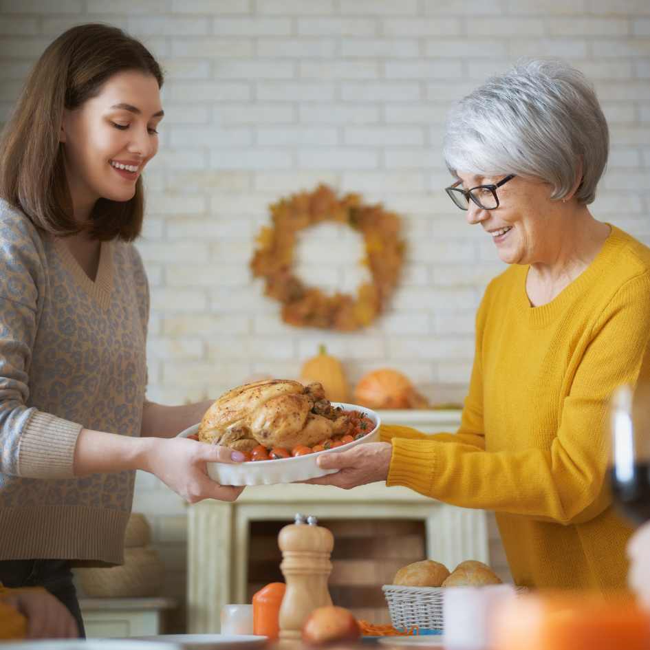 A woman hands a roasted turkey to an elderly woman in a festive, autumn-themed kitchen