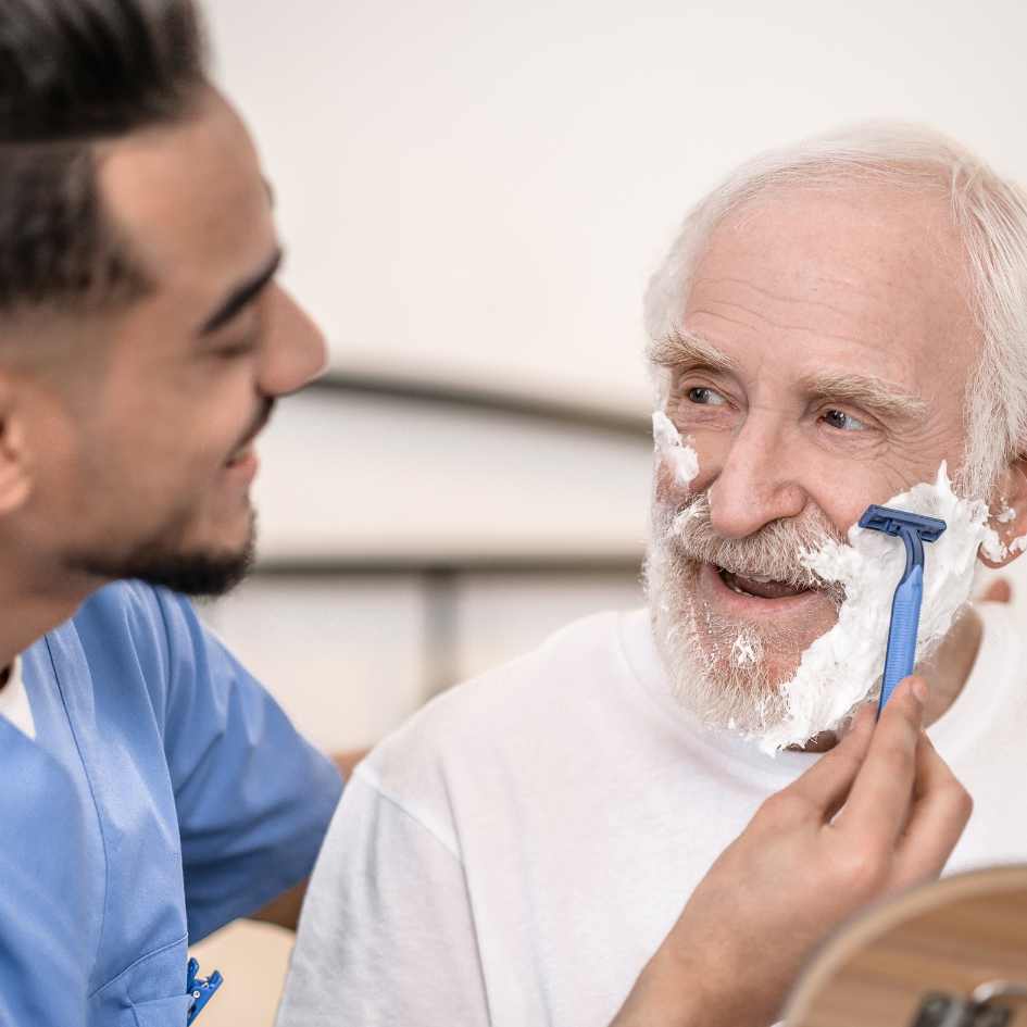 A caregiver helps an elderly man shave, both smiling during the interaction.