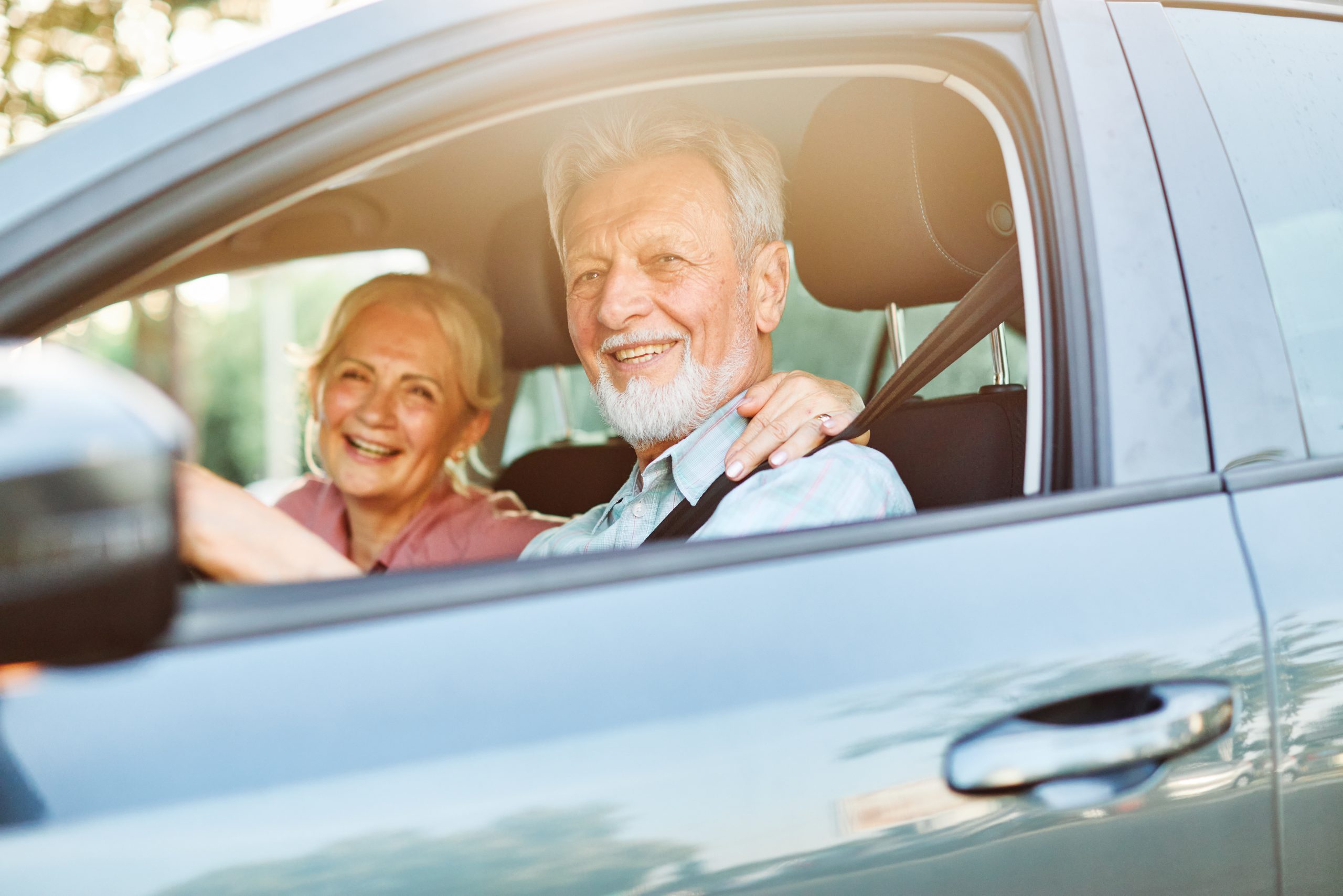 An older man in the drivers seat of a car wearing a seatbelt with an older woman sitting next to him.