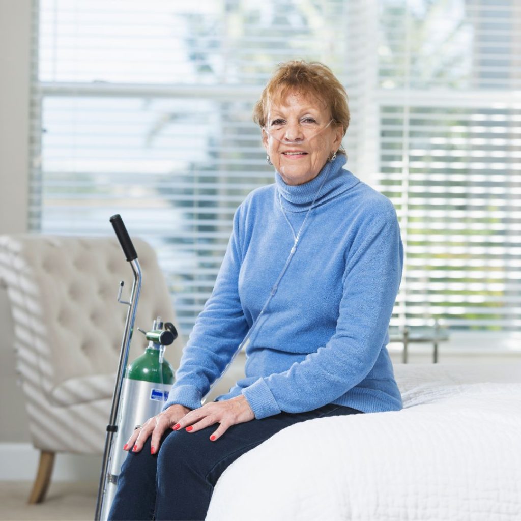 An elderly woman wearing a blue sweater, seated indoors with an oxygen tank smiling at the camera.