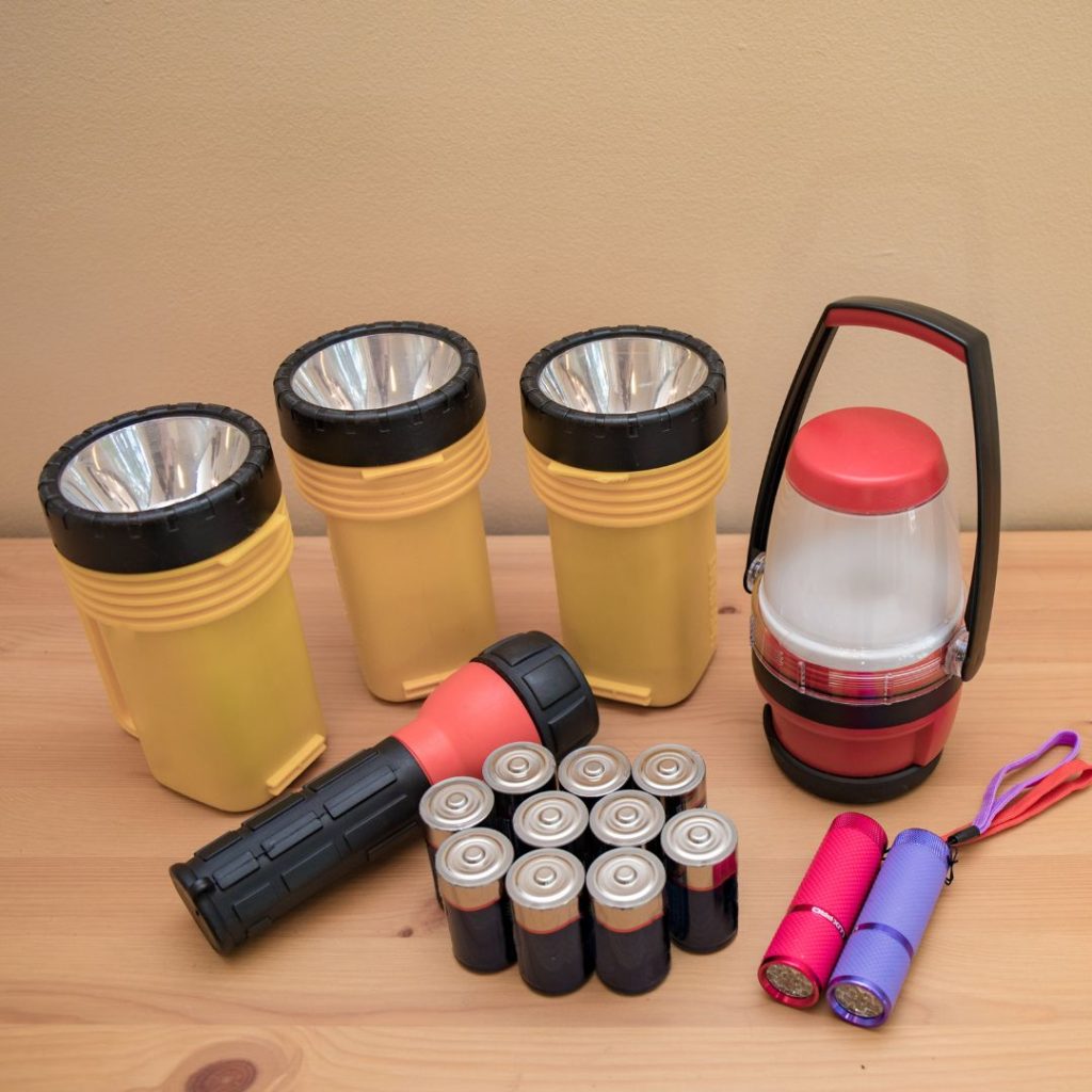 A variety of flashlights and batteries on a wooden surface, including large yellow flashlights, a lantern-style light, and two small colored flashlights.