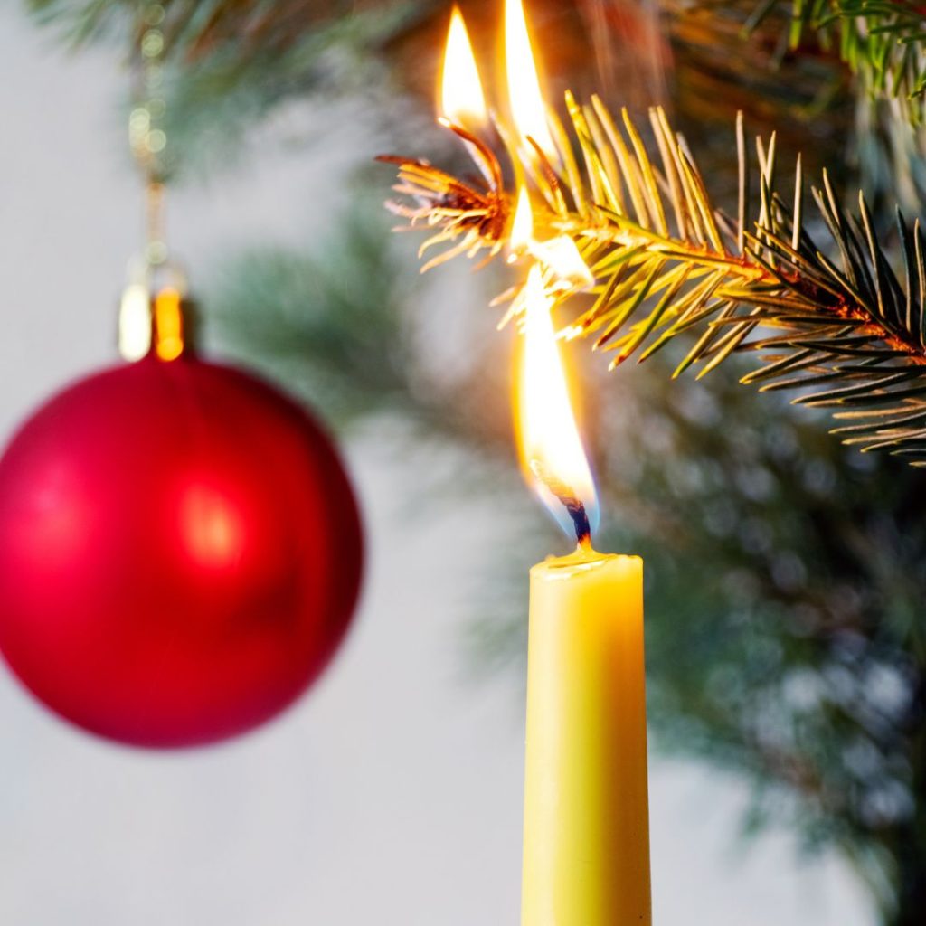 Up close shot of a lit candle catching a christmas tree branch on fire. Red ornament in background.