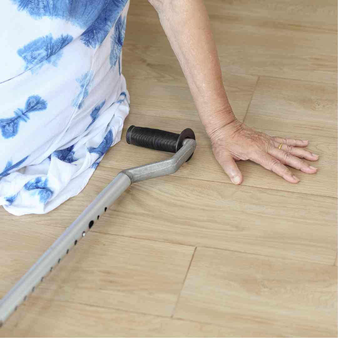 An older adult sitting on the floor, using their hand for support, next to a fallen cane, indicating a possible fall or difficulty getting up.