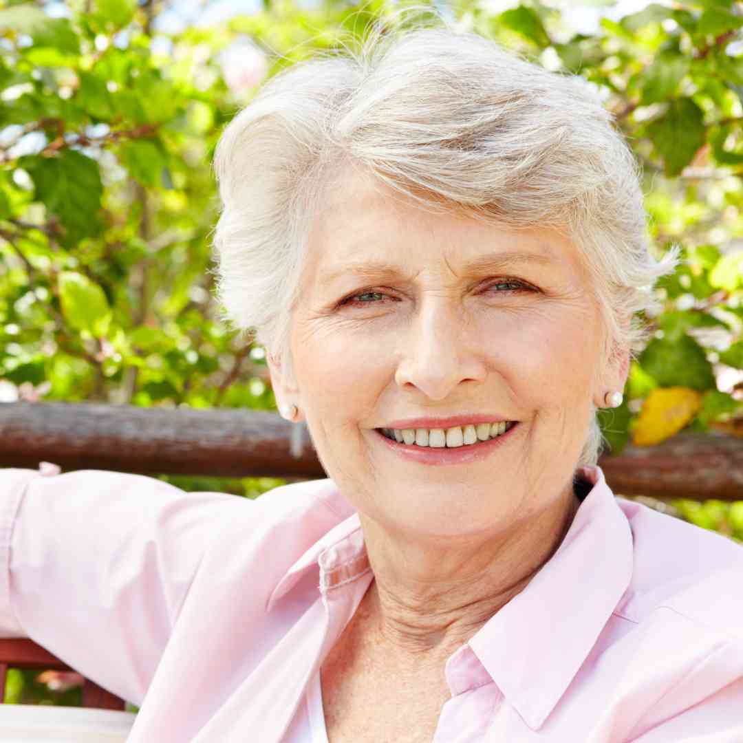 A smiling older woman with short gray hair sitting outdoors, wearing a light pink shirt, with lush greenery in the background.