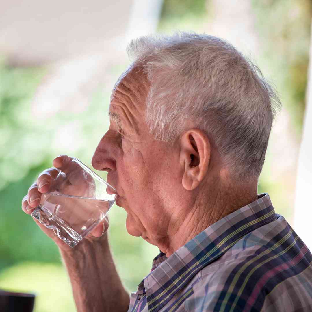 An older man with short gray hair drinking a glass of water, sitting outside, with blurred greenery in the background.