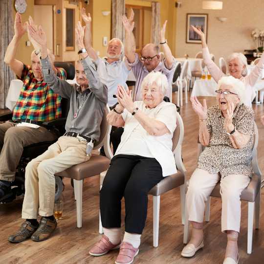 A group of older adults seated in chairs at a senior center, raising their arms and clapping together, participating in an interactive activity.