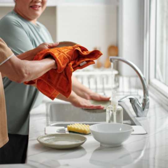 Two people washing and drying dishes at a kitchen sink, sharing a task with warm smiles and an orange dish towel