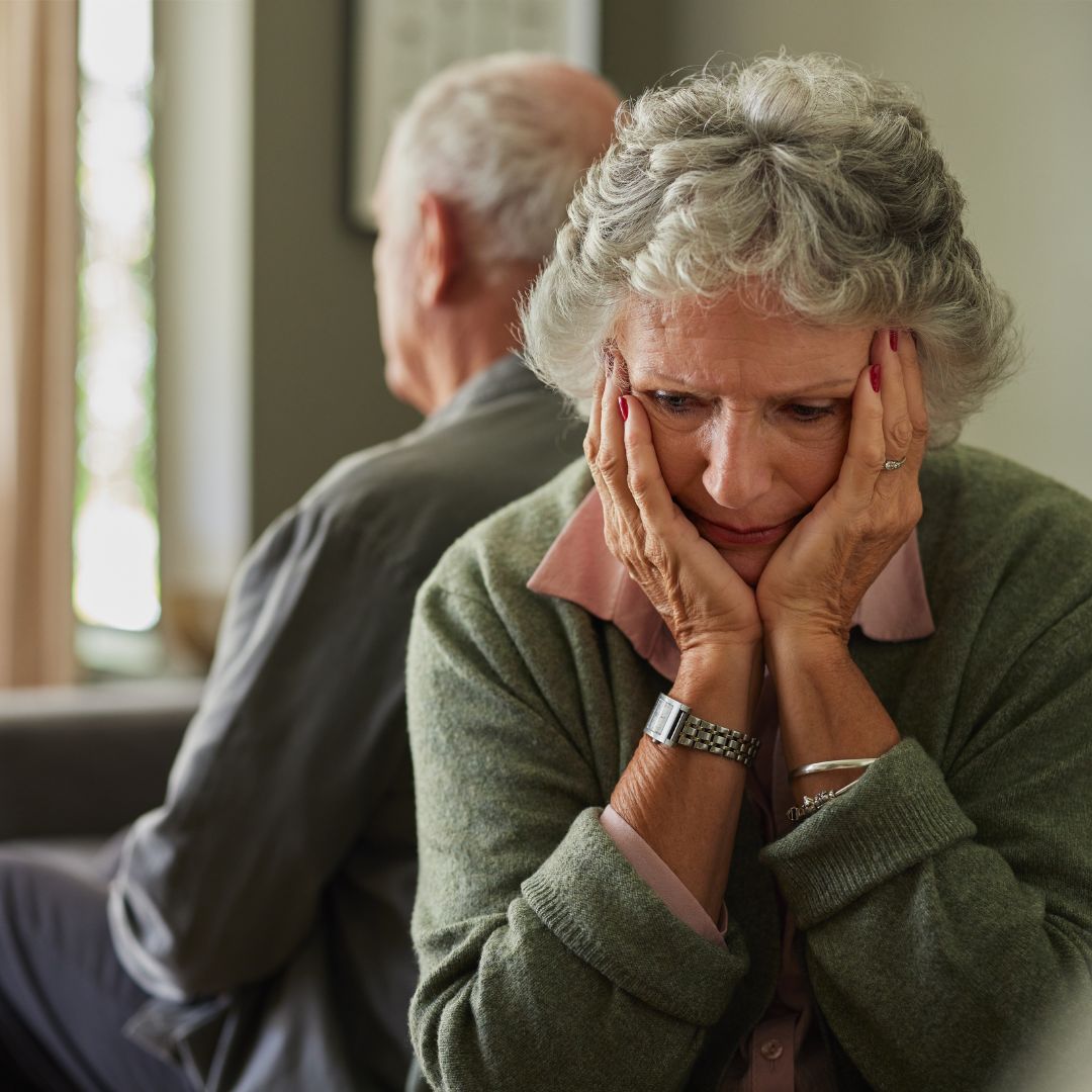 Older woman sitting with her head in her hands, looking upset, while an older man faces away in the background.