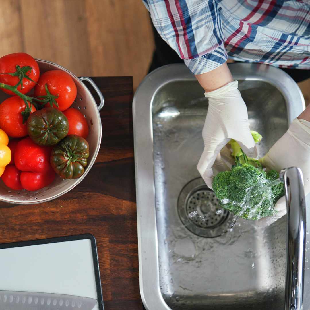 Person wearing gloves washing broccoli at a kitchen sink next to a bowl of fresh tomatoes and peppers.