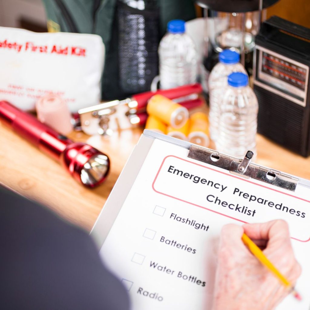 Hand filling out emergency checklist with supplies in background