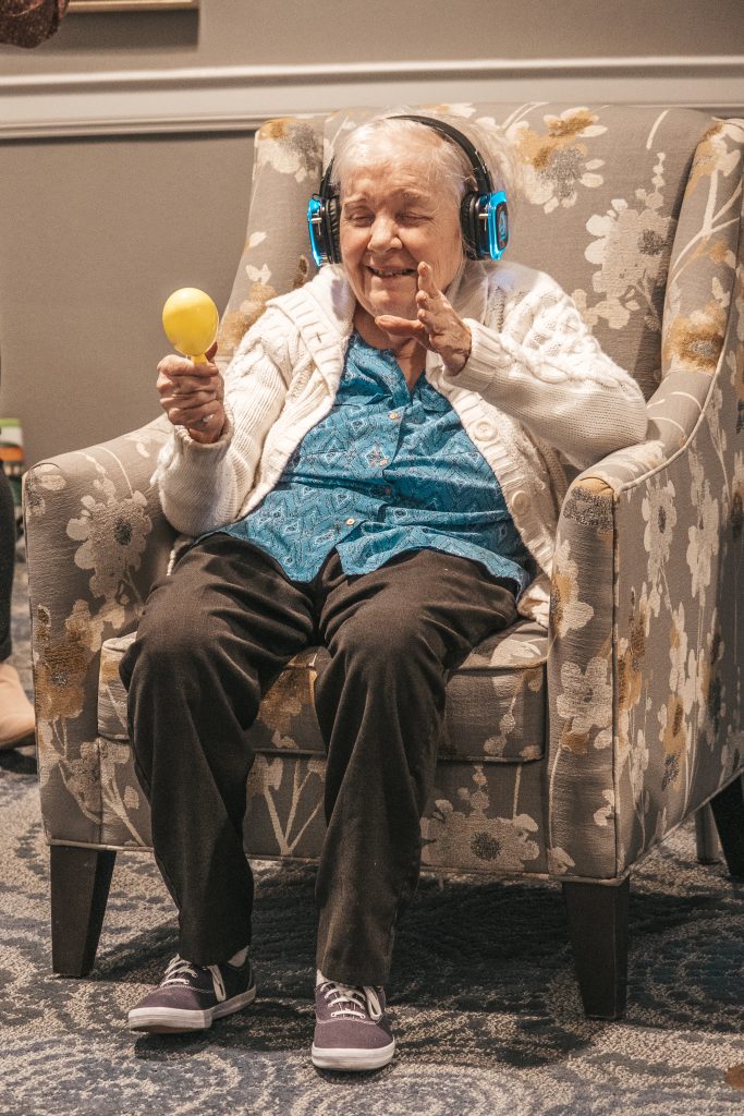 Elderly woman sitting in a floral chair wearing headphones, smiling with eyes closed, and holding a yellow maraca.