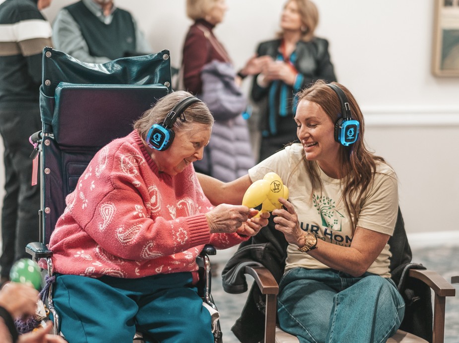 Elderly woman in a wheelchair wearing headphones, smiling while holding a yellow maraca with the assistance of a younger woman also wearing headphones.
