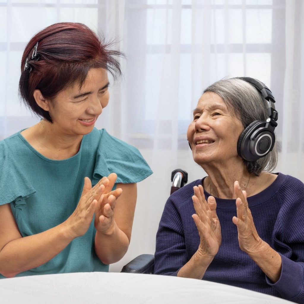 An elderly woman wearing headphones claps hands with a younger woman suggestive of music therapy.