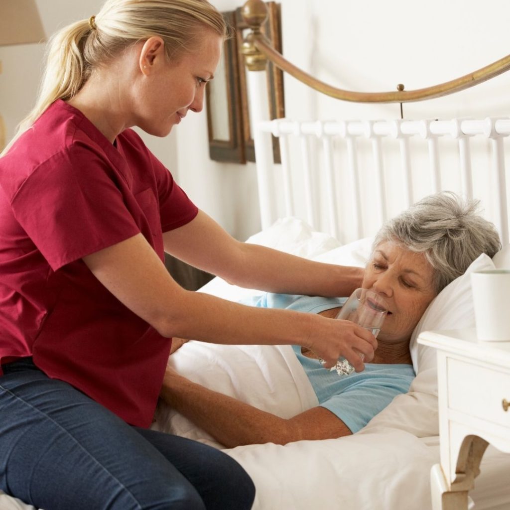 A caregiver assists an elderly woman in a bed by offering her a glass of water.