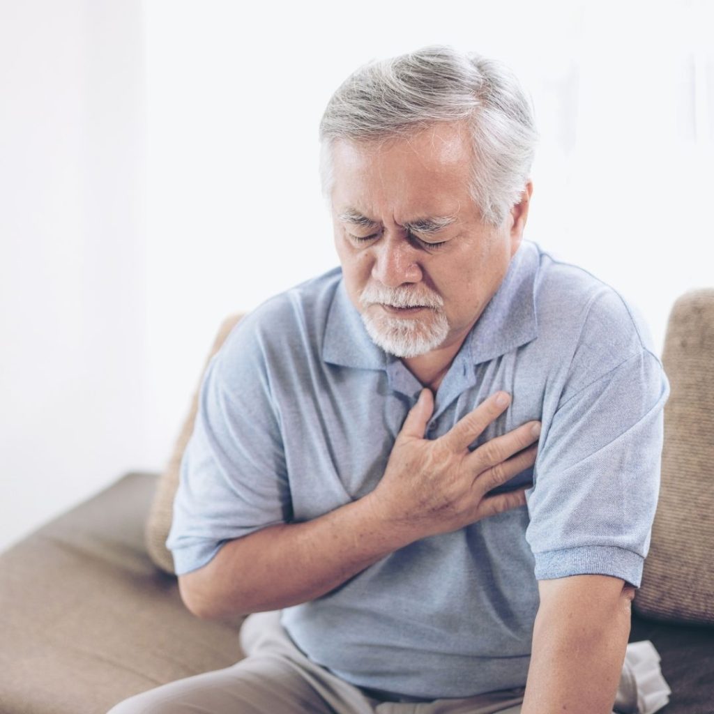 Older man sitting indoors, hand on chest, appearing to feel discomfort.