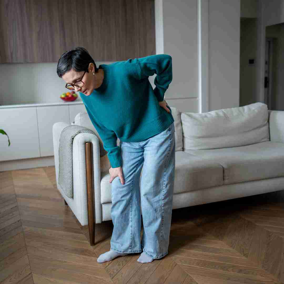Person standing beside a couch and holding their lower back and leg, suggesting back or joint pain.