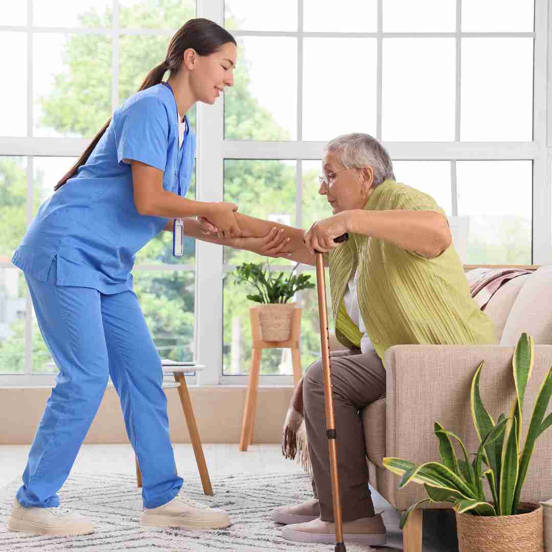Caregiver in blue scrubs helping an older adult stand up from a chair while using a cane.