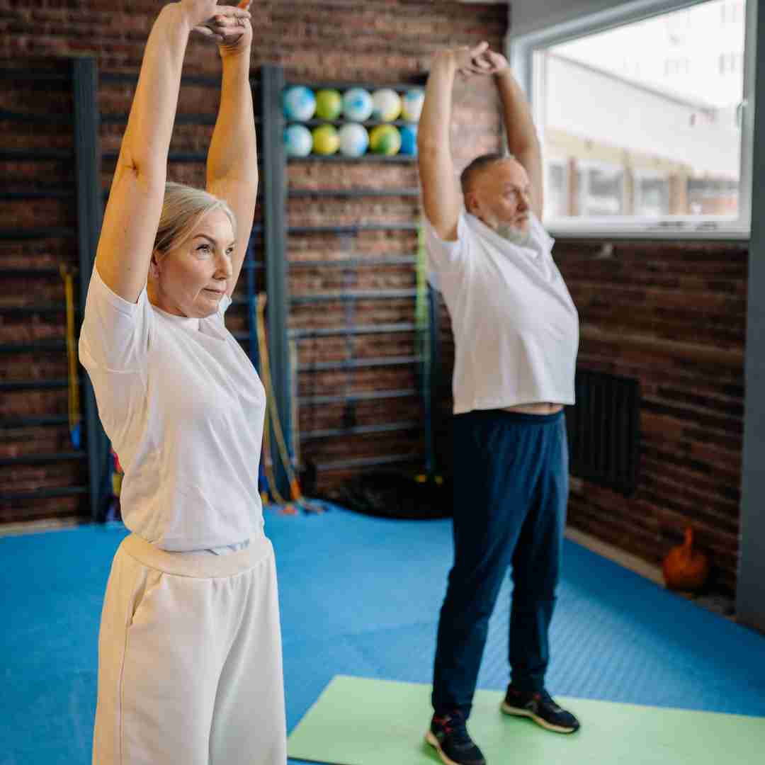 Two older adults in a fitness studio stretching with their arms raised overhead during exercise.