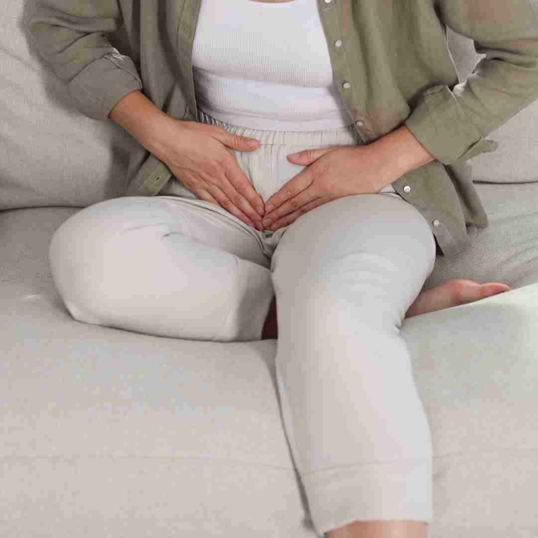 Person sitting on a couch with both hands placed over their lower abdomen, suggesting stomach pain or discomfort.