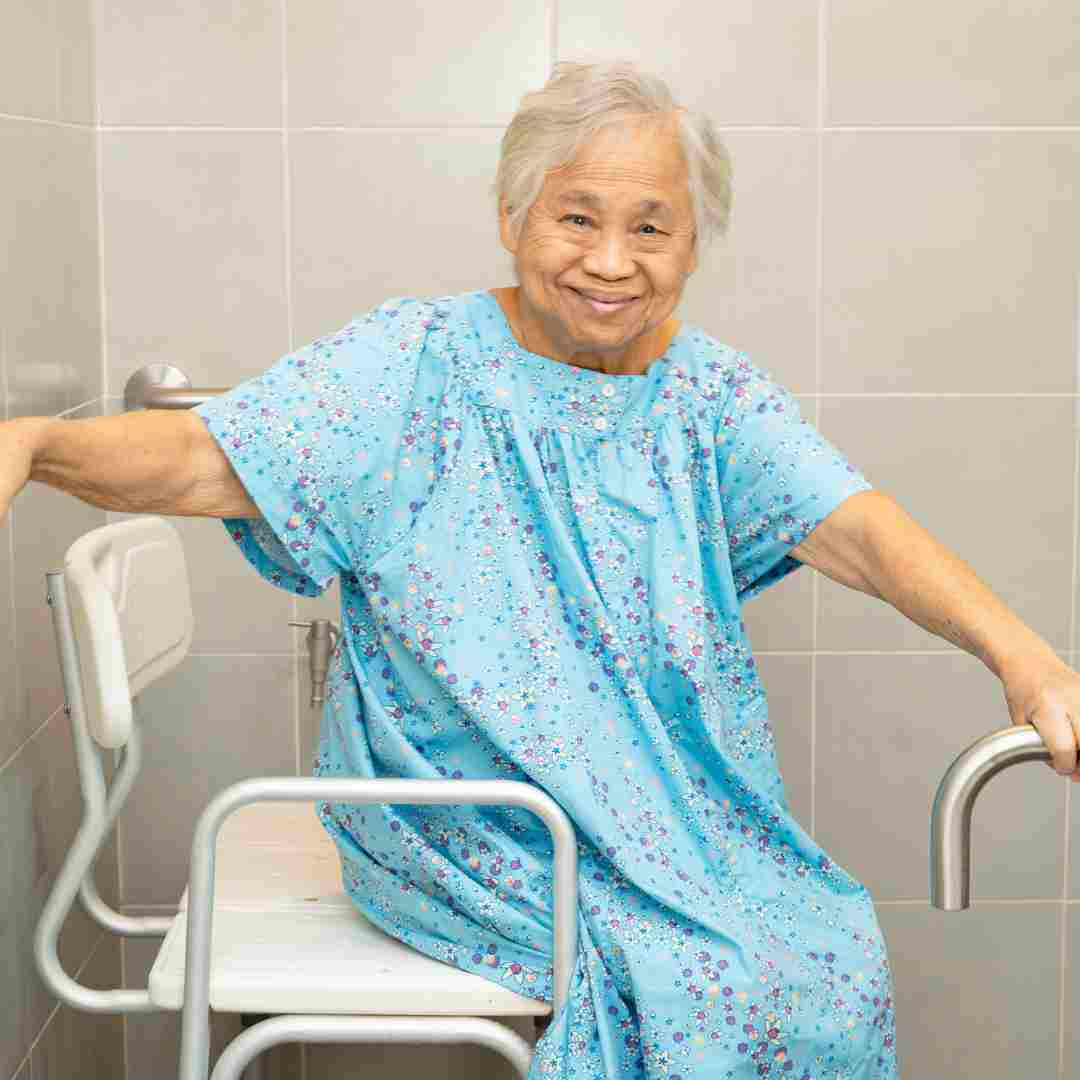 Older woman smiling while seated on a shower chair in a bathroom, holding grab bars for support.