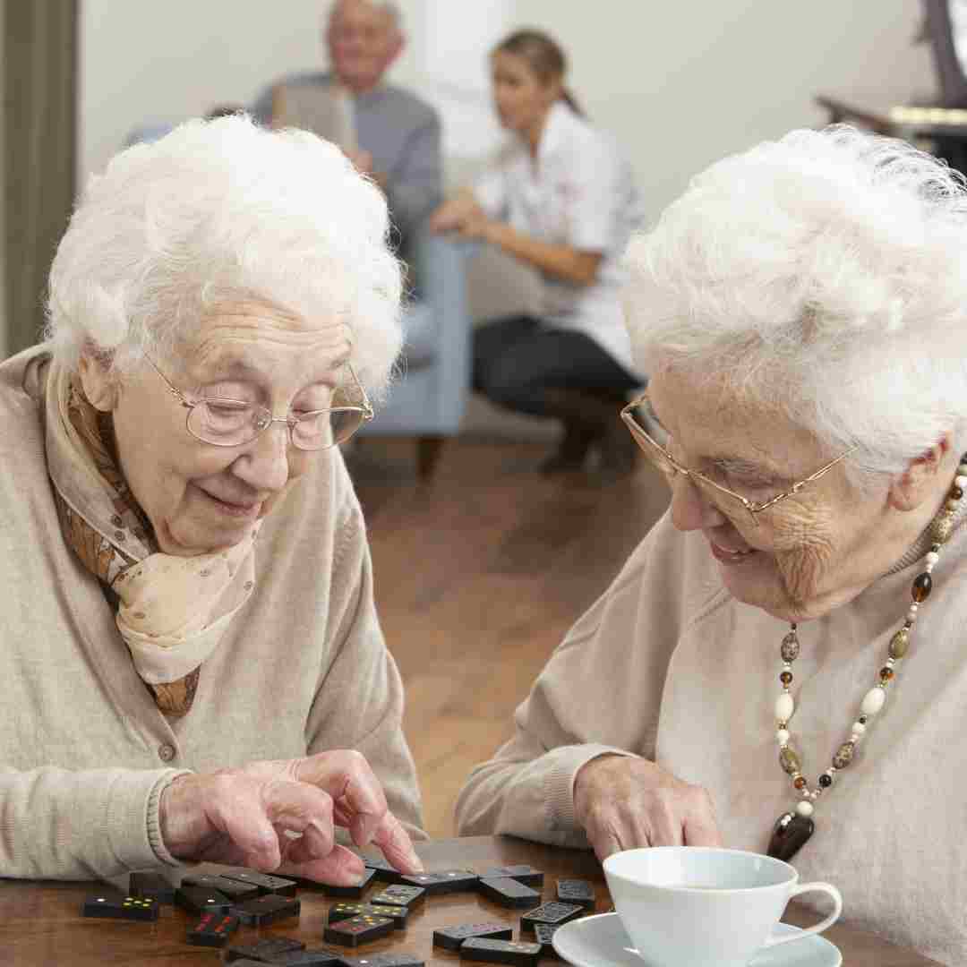 Two older women sitting at a table playing dominoes, with a caregiver and another older adult blurred in the background.