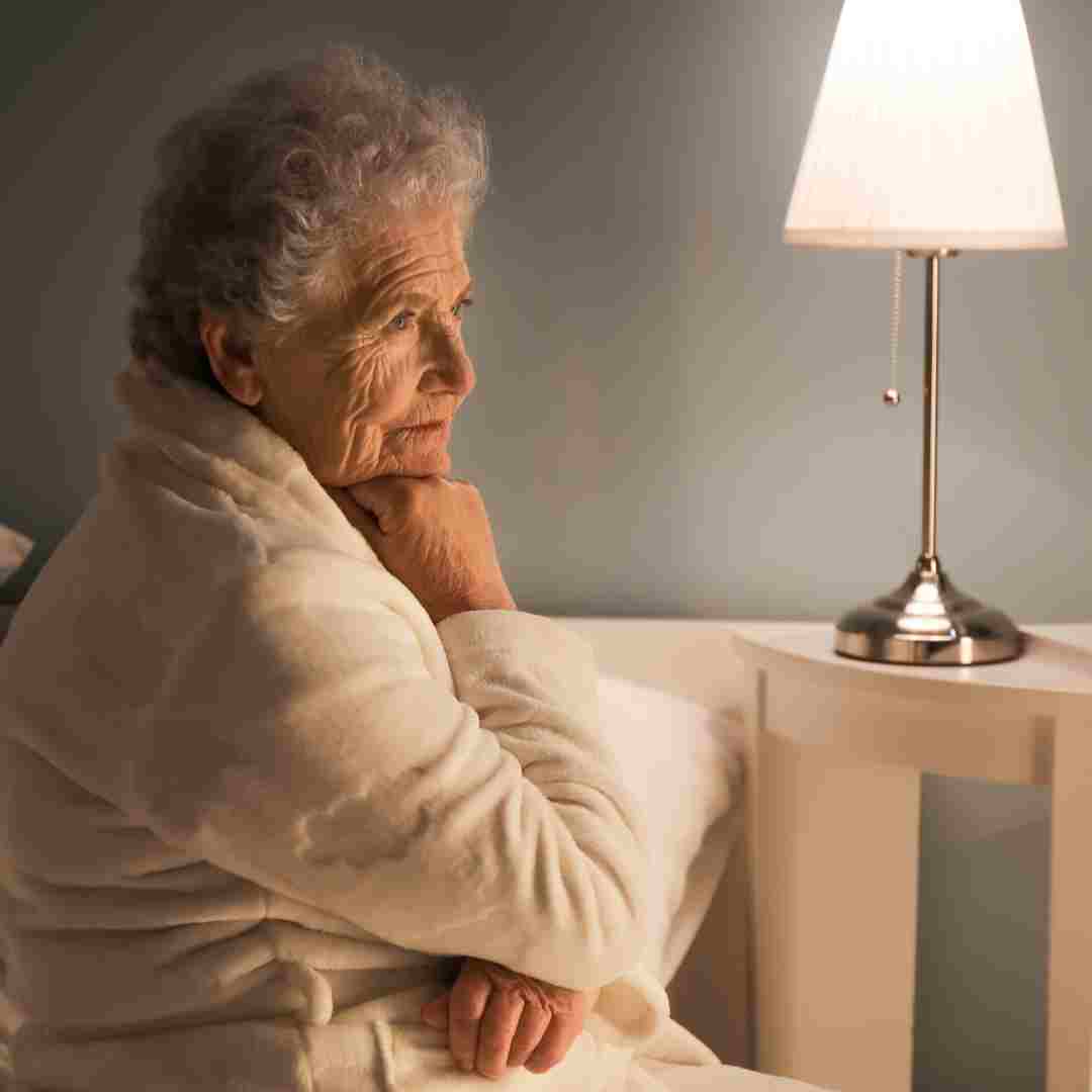 Older woman in a bathrobe sitting on a bed beside a lit table lamp.