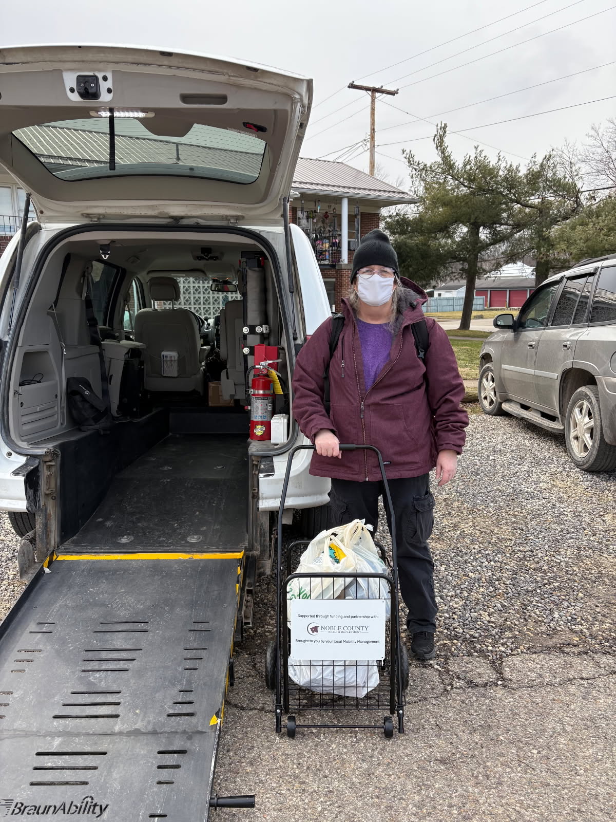 Person standing beside an accessible white van with the rear ramp lowered, holding a rolling grocery cart filled with bags in a parking area.