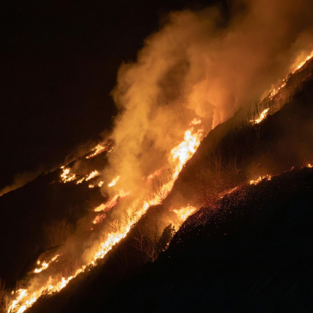 Wildfire burning across a hillside at night with heavy smoke and visible flames.