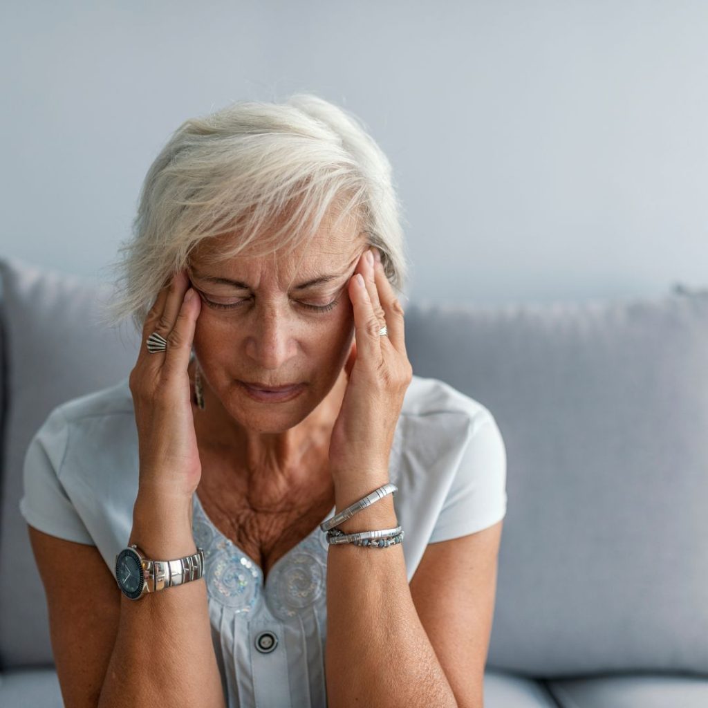 Older woman sitting on a couch holding her head, appearing stressed or in pain
