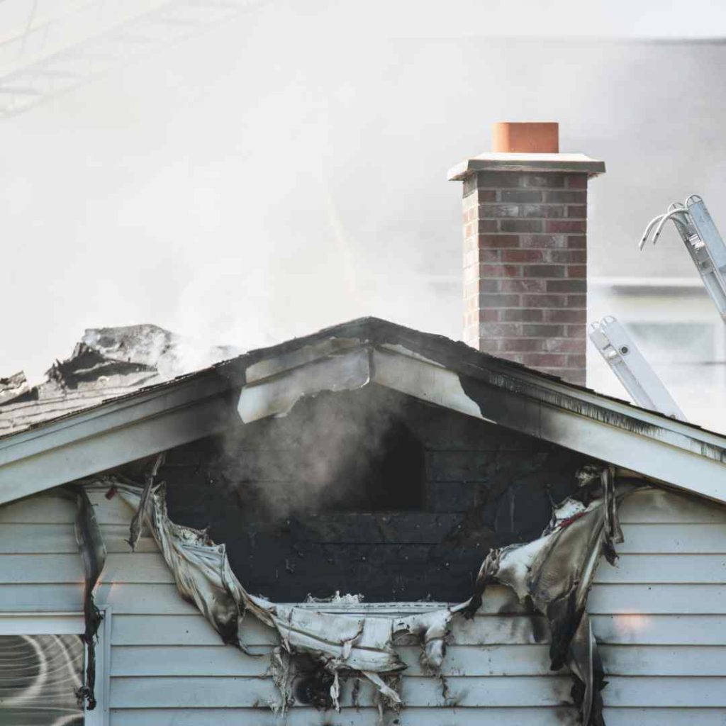 Smoke rises from a fire-damaged house with burned siding and roof damage.
