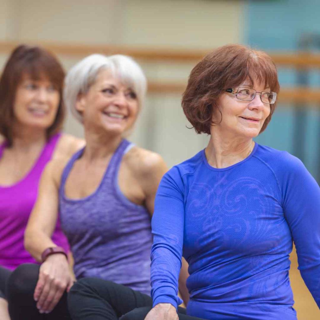 Three older women in exercise clothing sit together during a fitness class.