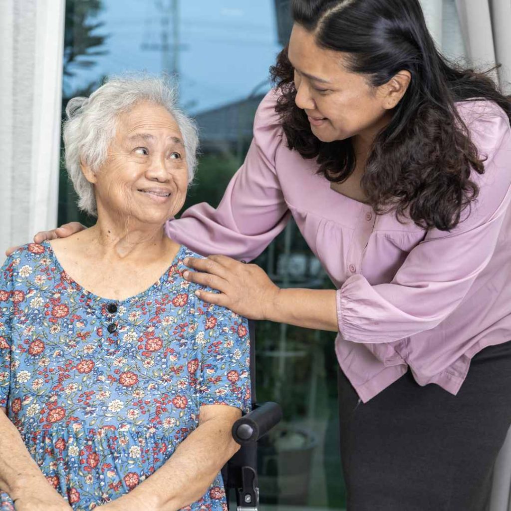 Older woman smiling with a caregiver standing beside her with a hand on her shoulder.