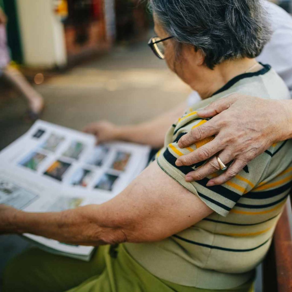 Older woman seated while looking at a photo album, with a hand resting on her shoulder.