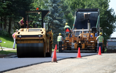 Road construction workers operate paving equipment while laying fresh asphalt on a roadway, with traffic cones marking the work area.