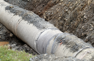 A large concrete pipe exposed in a dirt trench, with soil and gravel surrounding the pipe and grass visible in the foreground.