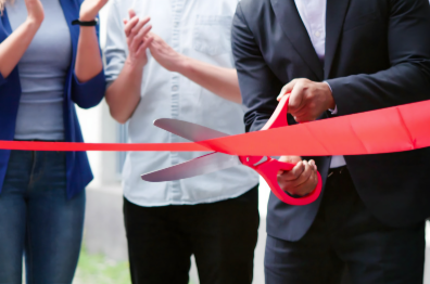 Person in a suit cuts a red ceremonial ribbon with oversized scissors as attendees applaud.