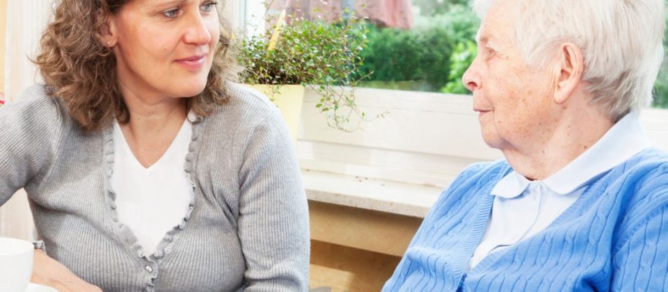 A woman in a gray cardigan listens to an elderly woman in a blue sweater over coffee.