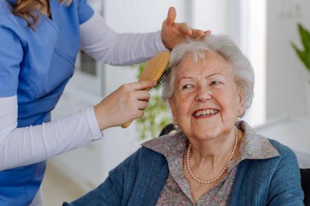 A woman helps an elderly woman who's sitting down brush her hair.