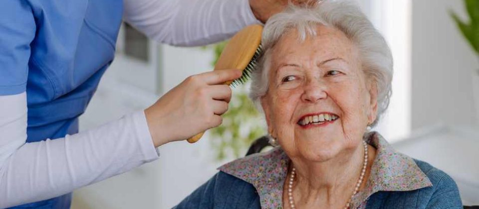 A woman helps an elderly woman who's sitting down brush her hair.