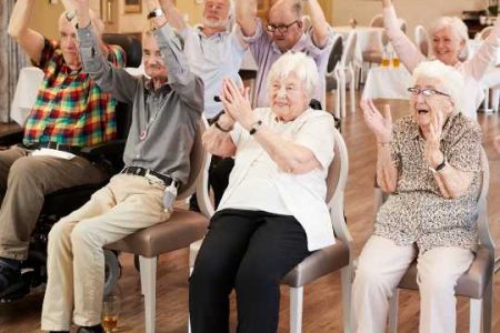 A group of older adults seated in chairs at a senior center, raising their arms and clapping together, participating in an interactive activity.